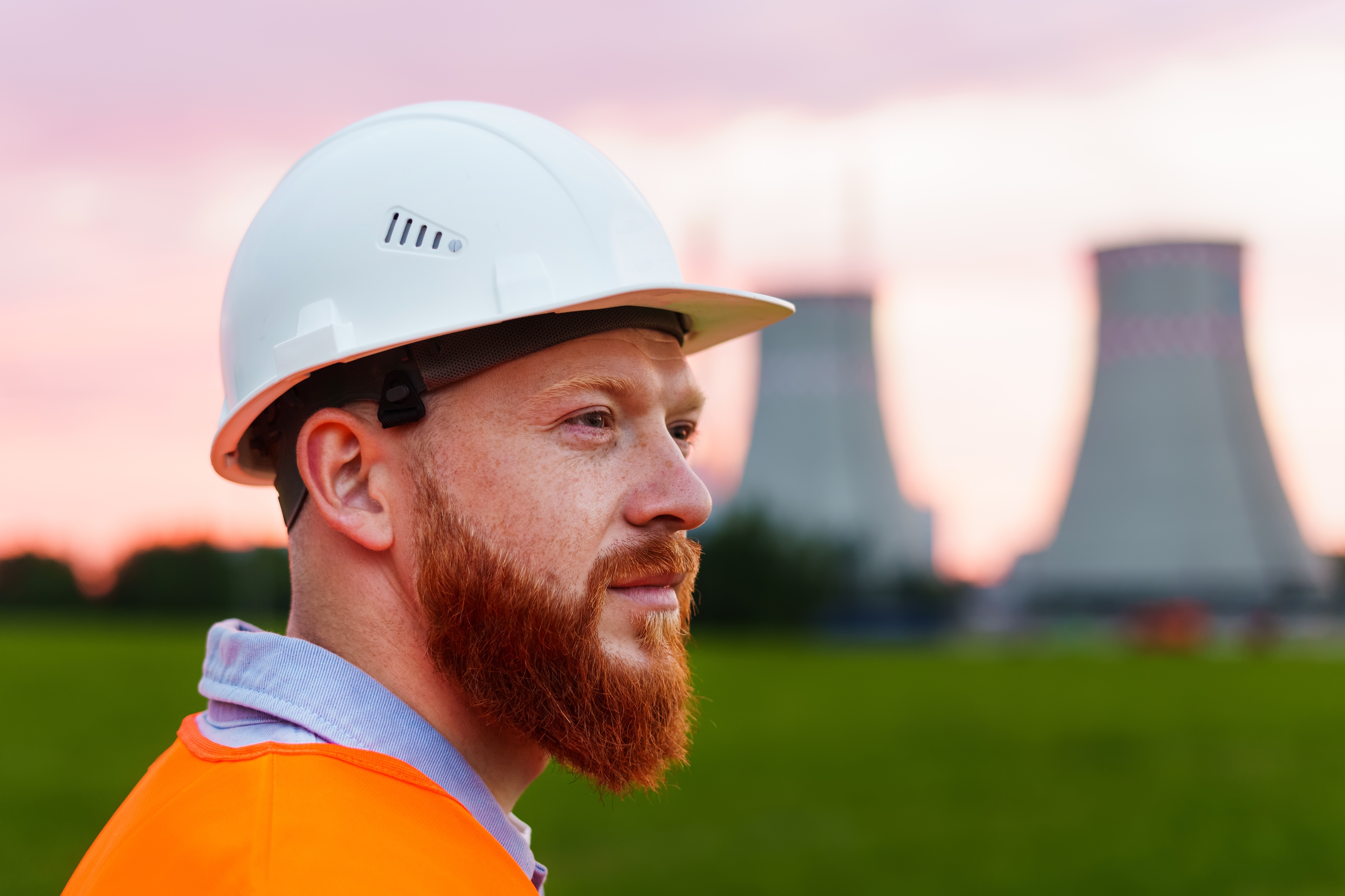 nuclear facility in background with white male in foreground wearing white hard hat, orange shirt, and having red beard.